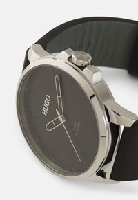 Crni sat s kućištem od srebrnog nehrđajućeg čelika, minimalističkim brojčanikom i crnim kožnim remenom. Brand "HUGO" istaknuto je prikazan.