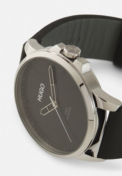 Montre noire avec un boîtier en acier inoxydable argenté, un cadran minimaliste et un bracelet en cuir noir. La marque "HUGO" est affichée de manière proéminente.