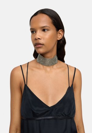 Metaliczny naszyjnik z teksturowanym siatkowym wzorem, o szerokiej szerokości i subtelnym połysku, noszony na czarnej satynowej sukience.