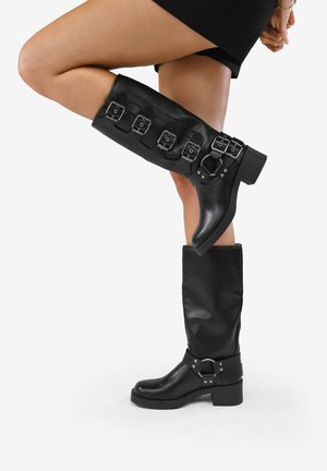 Femme portant des bottes en cuir noires montant jusqu'aux genoux avec plusieurs boucles argentées, une jambe levée et pliée sur un fond blanc uni.