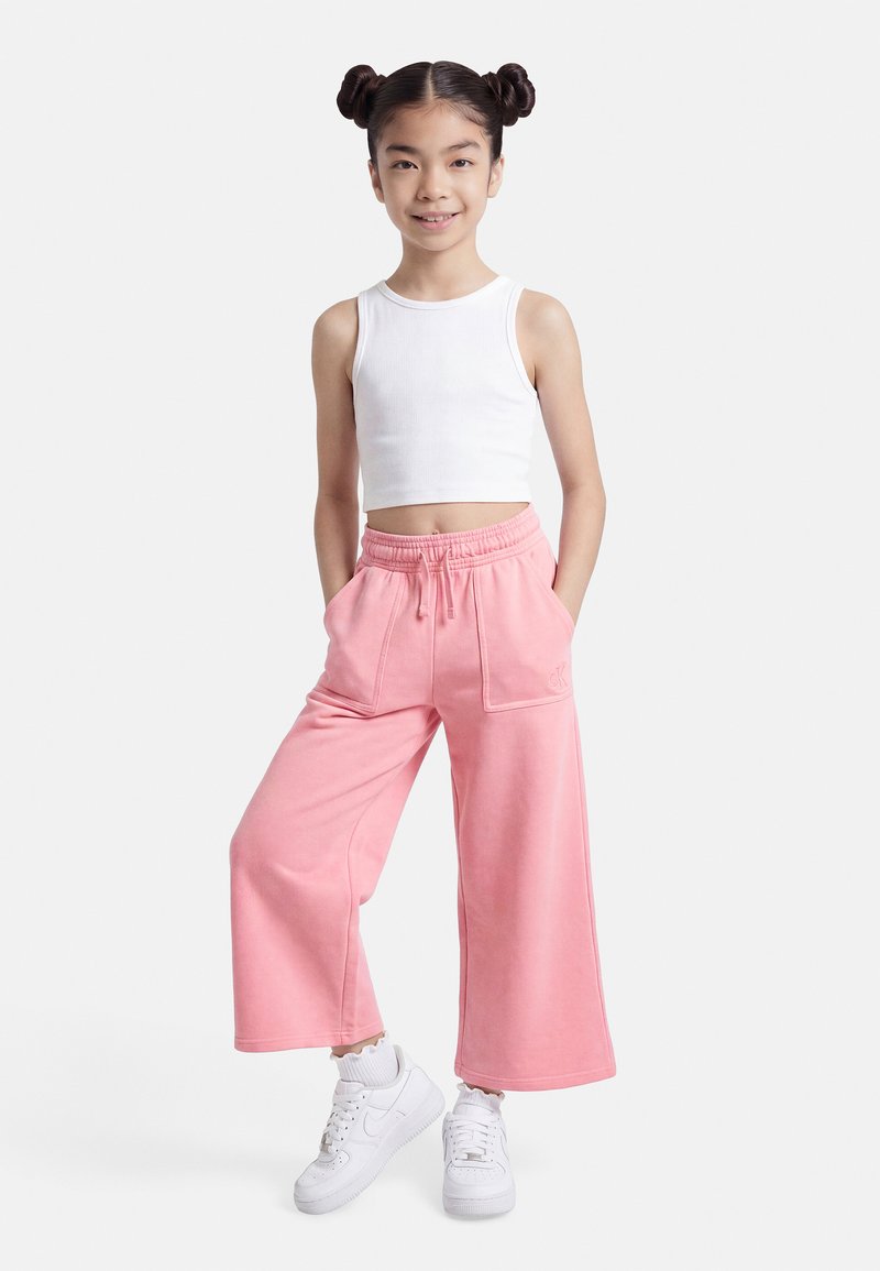 Νεαρή κοπέλα στέκεται με τα χέρια στις τσέπες φορώντας λευκό τοπ, ροζ φαρδιά παντελόνια, λευκές κάλτσες και λευκά αθλητικά παπούτσια, μπροστά σε απλό φόντο.