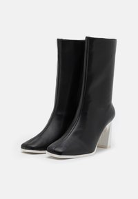 Botas de tobillo de cuero sintético negro con un alto tacón cuadrado y un diseño de puntera puntiaguda distintivo. Presentan un acabado suave y un acento de suela blanca.