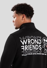 Sudadera negra con cuello alto. Gran texto y gráfico en la parte posterior, que muestra la frase "AMIGOS EQUIVOCADOS" y un personaje estilizado.
