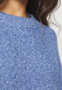 Pull en laine bleu tricoté avec un encolure ronde et un tissu texturé, présentant un motif chiné subtil. Gros plan sur le matériau et la forme.