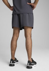 Γκρι αθλητικό σορτς από ελαφρύ ύφασμα, με άνετη εφαρμογή. Μαύρα αθλητικά παπούτσια με λευκές λεπτομέρειες και λογότυπο στη φτέρνα.