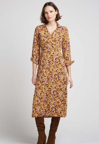 Robe à manches longues, fleurie, de couleur jaune moutarde avec des accents violets et blancs. Avec un col, une taille cintrée et une longueur mi-mollet. Portée avec des bottes marron.