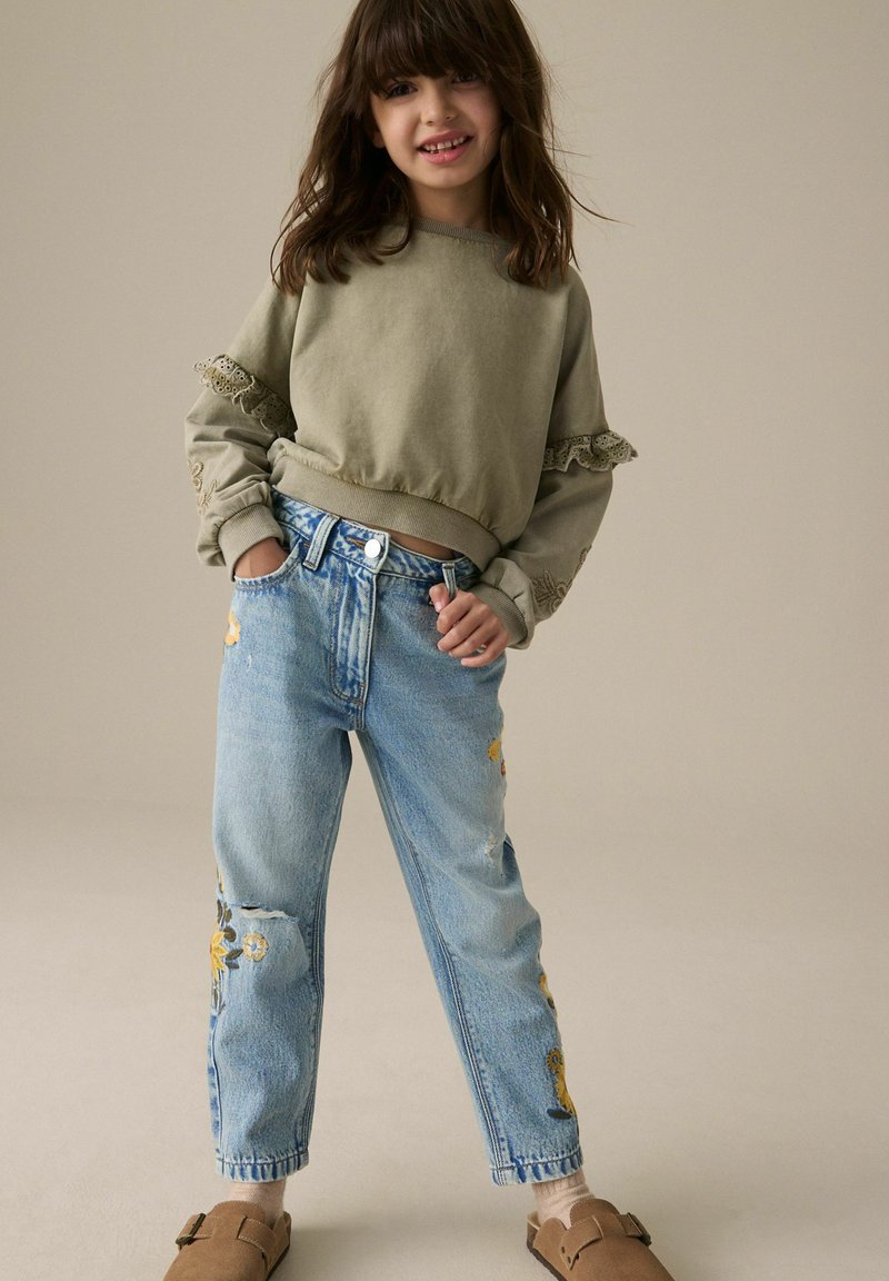 Ung pige med brunt hår iført en grøn sweatshirt med blondedetaljer, blå blomsterbroderede jeans og brune sko med spænder, stående med hænderne i lommerne.