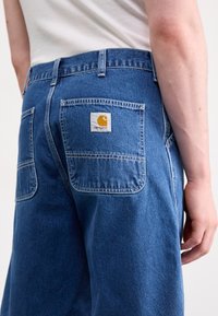 Tamsiai mėlyni džinsiniai šortai, turintys užpakalines kišenes su horizontaliu siuvimu ir Carhartt logotipo ženkliuku dešinėje kišenėje.