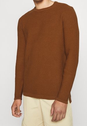 Homme portant un pull en tricot brun texturé à manches longues et un short de couleur crème, debout devant un fond blanc uni.