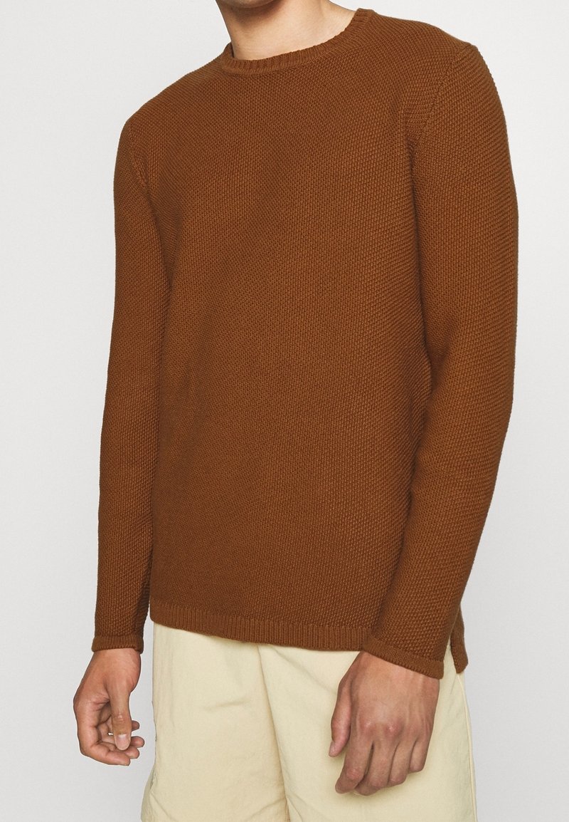 Homme portant un pull en tricot brun texturé à manches longues et un short de couleur crème, debout devant un fond blanc uni.