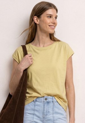 Ung kvinde i gul kortærmet skjorte og lyseblå jeans holder en brun skuldertaske, smiler og kigger mod højre mod en lys væg.