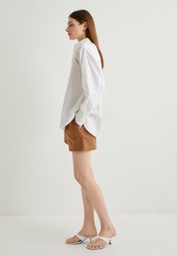 Hvid linned skjorte med knapper og en løs pasform, brune skræddersyede shorts og hvide hælsandaler, der fremhæver en minimalistisk stil.