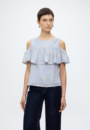 Γυναίκα με ανοιχτό μπλε μπλούζα με ασημένιο σχέδιο, με ακάλυπτους ώμους και βολάν, και σκούρο φαρδύ παντελόνι, στέκεται μπροστά σε απλό λευκό φόντο.