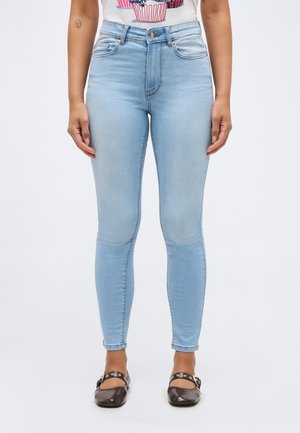 Nő világoskék skinny farmerben, egy cupcake mintás fehér felsőben és sötétbarna Mary Jane cipőben ezüst szemes díszítéssel.