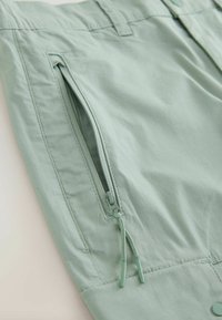 Section de tissu vert clair avec une poche verticale à fermeture éclair partiellement ouverte et double curseurs de fermeture éclair avec extrémités cordées.