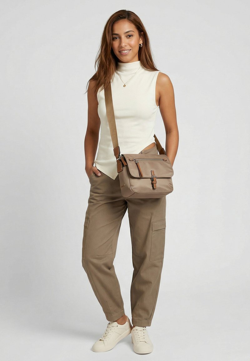 Femme debout souriante, portant un haut blanc sans manches, un pantalon cargo kaki, des baskets blanches et un sac bandoulière beige.