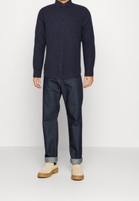 Camicia blu navy con maniche lunghe, una tasca sul petto, abbinata a jeans in denim scuro e scarpe beige slip-on con suola spessa.