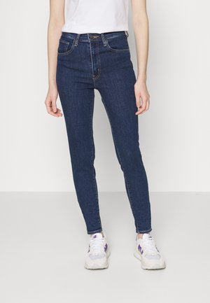 Personne portant un jean slim bleu foncé, des baskets blanches avec des accents violets, et un haut blanc à manches courtes, debout devant un fond uni.
