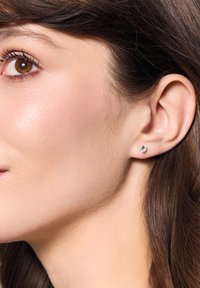 Okrągła srebrna kolczyk z przejrzystym kamieniem w centrum, osadzona w gładkiej metalowej bazie, prezentowana na uchu modelki na tle gładkiej skóry.