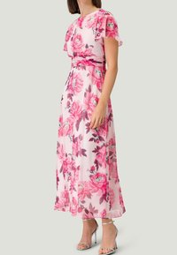 Robe maxi florale en tissu rose transparent avec des manches à volants. Présente des fleurs roses et vertes vives, une taille cintrée, et une jupe fluide.