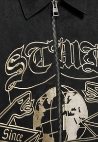 Čierna zipsová bunda s zlatou výšivkou globu a dekoratívnymi grafikami, s textúrovaným materiálom a špicatým golierom.