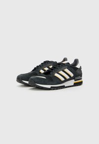 Black suede sneakers with mesh accents, featuring three white stripes, a white midsole, and a yellow and black heel. Round toe and lace-up design.
