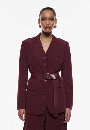 Blazer tailored bordeaux con maniche lunghe, chiusura a un bottone, indossato su un top abbinato, segnato in vita con una cintura marrone.