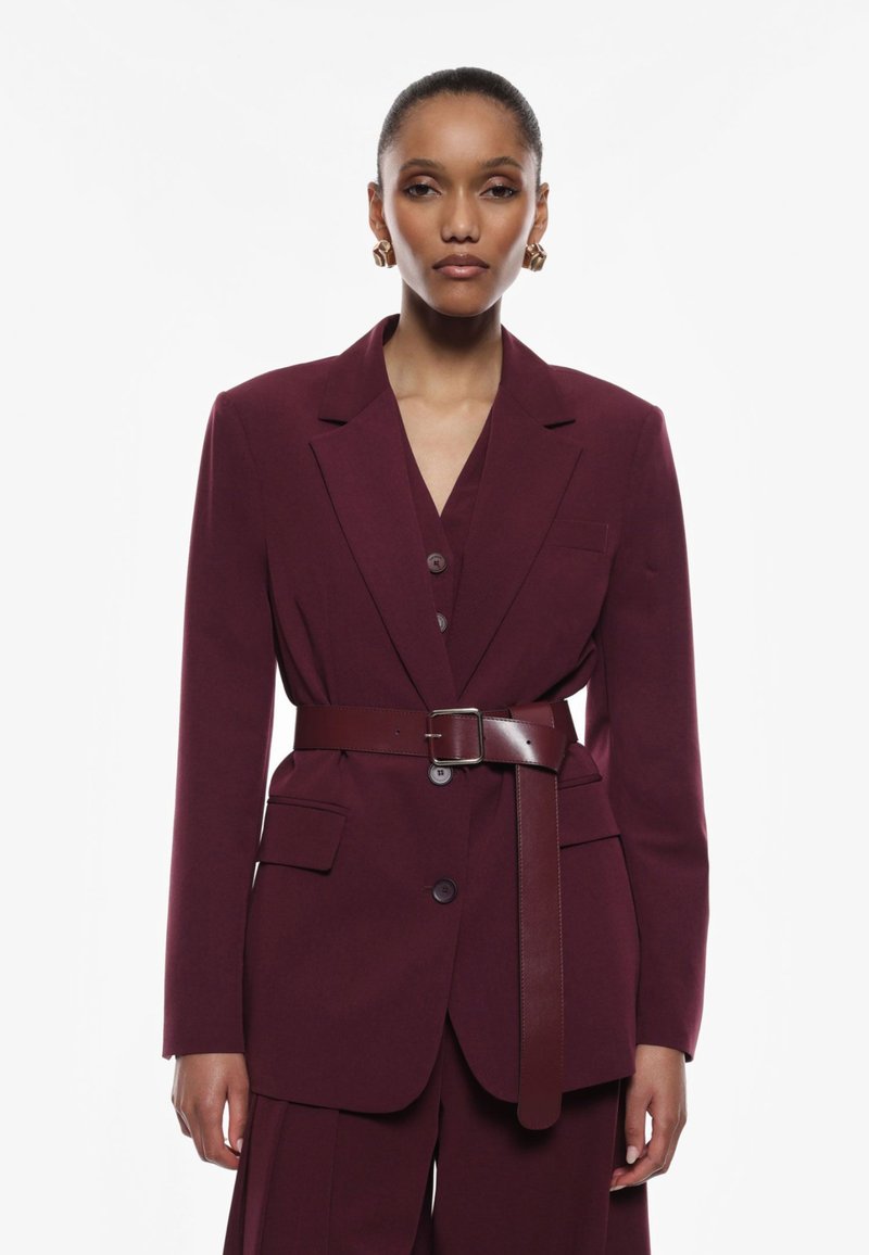 Blazer su misura color bordeaux con vita fasciante, bottoni monopetto e tasche frontali. Caratterizzato da un design strutturato e una texture di tessuto liscia.