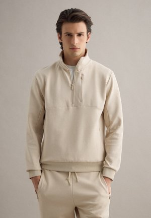 Junger Mann mit dunklen Haaren, der einen passenden beigen Pullover mit Viertel-Reißverschluss und beige Hosen mit Kordelzug trägt, stehend vor einem einfarbigen Hintergrund.