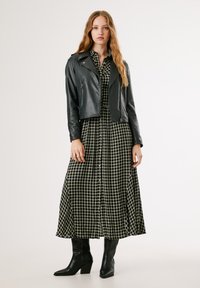 Casaco de motoqueiro em couro preto sobre um vestido longo com padrão às riscas em preto e branco. A modelo usa botas de salto pretas. Fundo minimalista.