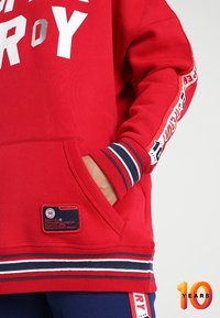 Rode sweatshirt met een groot zilveren logo, geribde manchetten met marineblauwe en witte strepen, en een merkpatch aan de onderzoom.