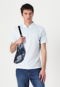 Svijetloplavi polo majica s ovratnikom na zip, uparen s tamnoplavim trapericama i malom torbom preko ramena u tie-dye stilu s detaljima na zip.