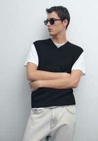 Maglione senza maniche a coste nere sopra una t-shirt bianca, abbinato a jeans grigi chiari; il modello posa contro un muro neutro.