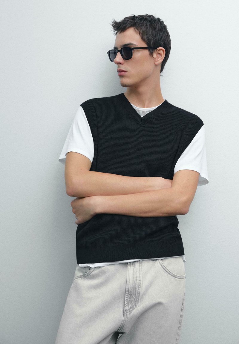 Maglione senza maniche a coste nere sopra una t-shirt bianca, abbinato a jeans grigi chiari; il modello posa contro un muro neutro.