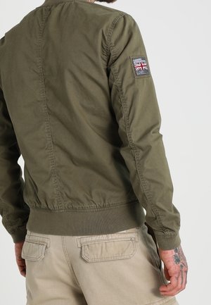 Mann trägt olivgrüne Jacke mit britischem Flaggen-Patch am Ärmel und beigefarbene Cargohosen, zeigt Rücken und Tätowierung an der rechten Hand.