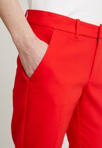 Un pantalon rouge sur mesure avec une texture lisse présente une poche latérale et un devant plat. La taille est structurée avec des passants pour ceinture.