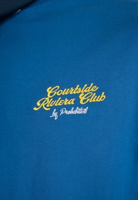 Tissu bleu avec texte brodé en jaune et blanc "Courtside Riviera Club by Prohibited" sur la région de la poitrine.