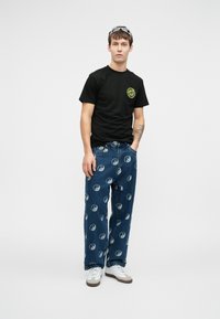 Νεαρός άνδρας που στέκεται φορώντας μαύρο μπλουζάκι Santa Cruz, μπλε τζιν με άσπρα σύμβολα yin-yang, λευκά αθλητικά παπούτσια και γυαλιά ηλίου στο κεφάλι.