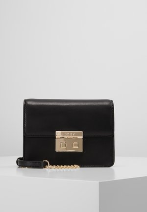 Bolso de mano de cuero negro con cierre rectangular en tono dorado y detalle de cadena, colocado sobre una superficie blanca frente a un fondo gris liso.