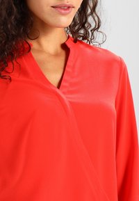 Blusa rossa con una texture liscia, scollatura a V e design avvolgente. Maniche lunghe e una leggera lucentezza mettono in risalto il materiale.