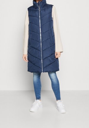 Person iført en ærmeløs, marineblå, quiltet lang vest med lynlås foran, beige bluse med lange ærmer, blå jeans og hvide sneakers.