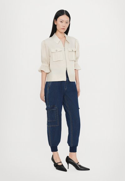 Beige zip-up jacket with two chest pockets and puffed sleeves, paired with dark blue cargo pants featuring pocket details and elastic cuffs. Black heels.