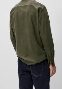 Homme portant une chemise à manches longues olive et un jean bleu foncé, vu de dos sur un fond clair et uni.