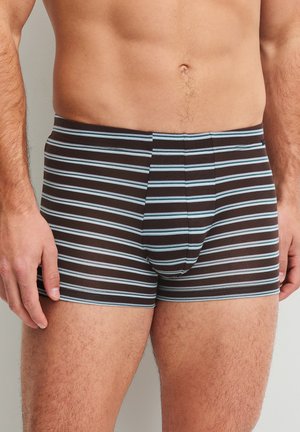 Torace maschile che indossa boxer aderenti con strisce orizzontali azzurro chiaro e bianche su sfondo scuro, in piedi contro una parete liscia.
