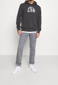 Felpa con cappuccio grigio scuro con una grafica frontale, abbinata a pantaloni grigi chiari arrotolati e sneaker bianchi. Composizione in misto cotone, vestibilità casual e texture liscia.
