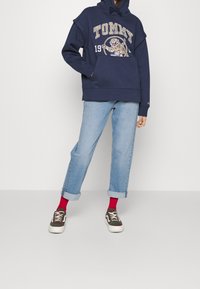 Sudadera navy con diseño de logo "TOMMY", bolsillo canguro, combinada con jeans azul claro enrollados y zapatillas con estampado acompañadas de calcetines rojos.