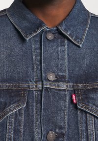 Chaqueta de mezclilla azul oscuro con cuello, cierres de botones metálicos y una etiqueta roja de Levi's en el costado. Cuenta con bolsillos estructurados y costuras.