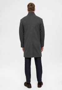 Grauer Wollmantel mit hohem Kragen, geradem Schnitt und Rücken-Schlitz, getragen über dunkelblauen Hosen und braunen Schuhen.