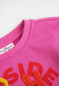 Różowa bawełniana bluza z dużym czerwonym napisem, teksturowanym puszystym pomarańczowym akcentem w kształcie królika oraz żebrowanym dekoltem typu crew. Widoczne metki.