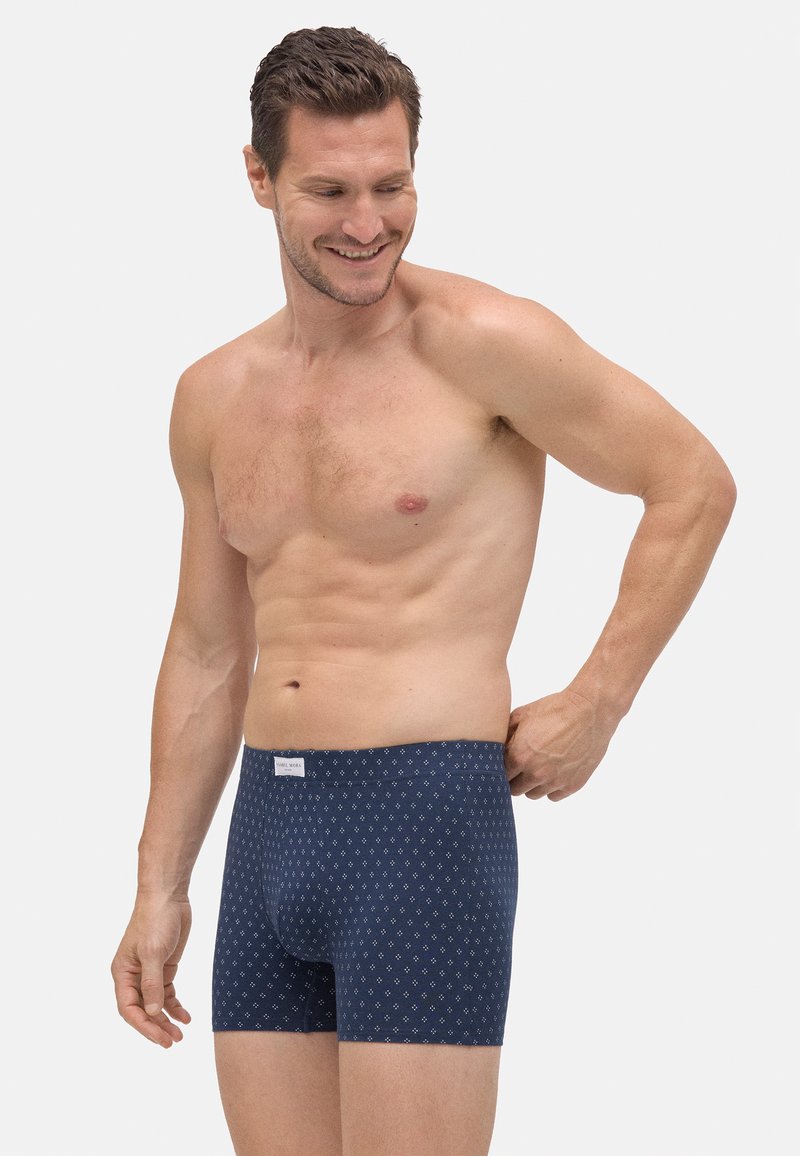 Hombre sonriendo sin camisa ajustándose un calzón azul marino con estampado contra un fondo blanco liso.